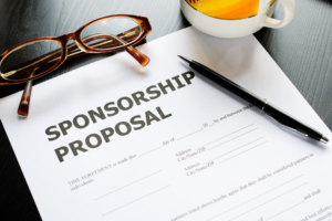 A sponsorship proposal rests on a desk by a pair of glasses, mug, and a black ballpoint pen.