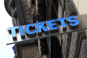 An upward angle shows a lighted, blue “Tickets” sign at a ticket booth outside an event venue.