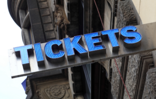 An upward angle shows a lighted, blue “Tickets” sign at a ticket booth outside an event venue.